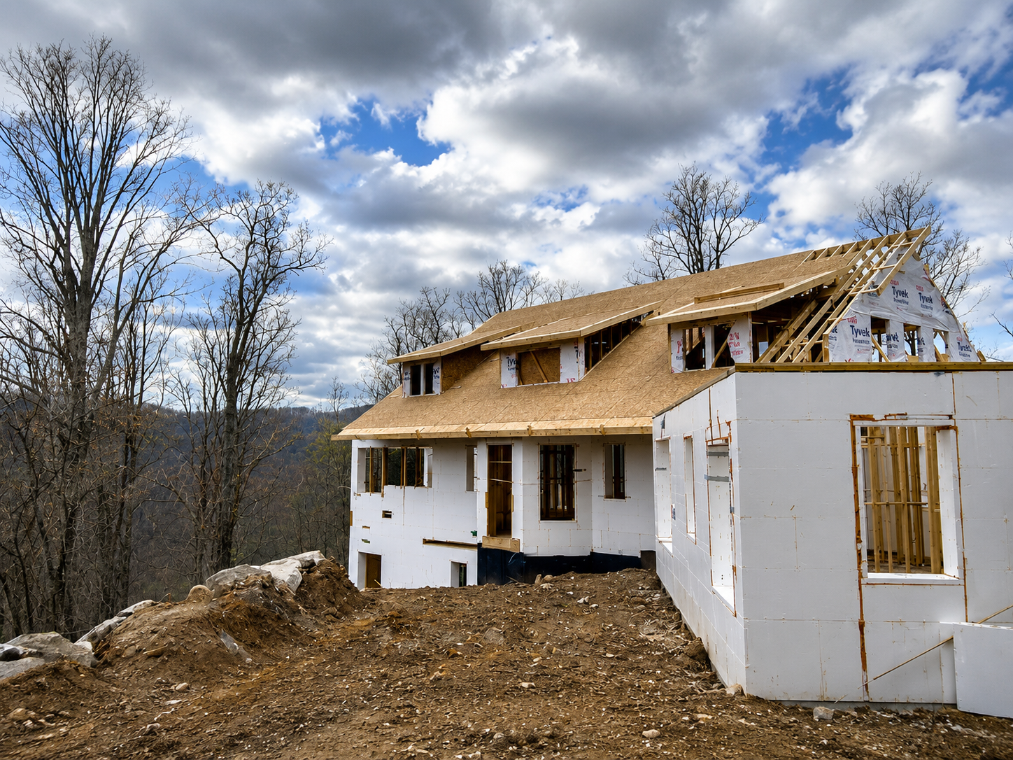 Partially built white house with exposed wooden roof framing on a wooded hillside under a cloudy sky