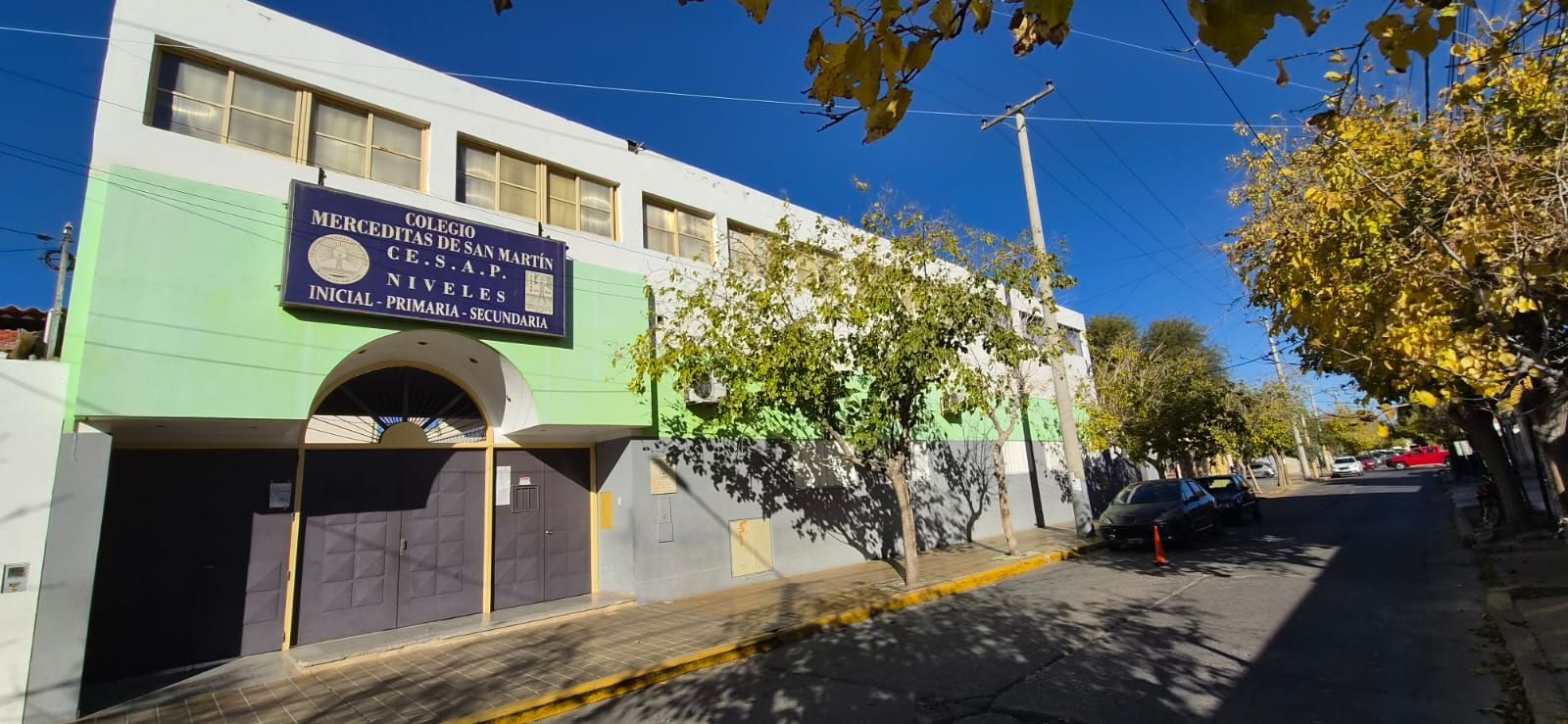Edificio escolar con fachada verde y letrero azul a lo largo de una calle soleada bordeada de árboles.
