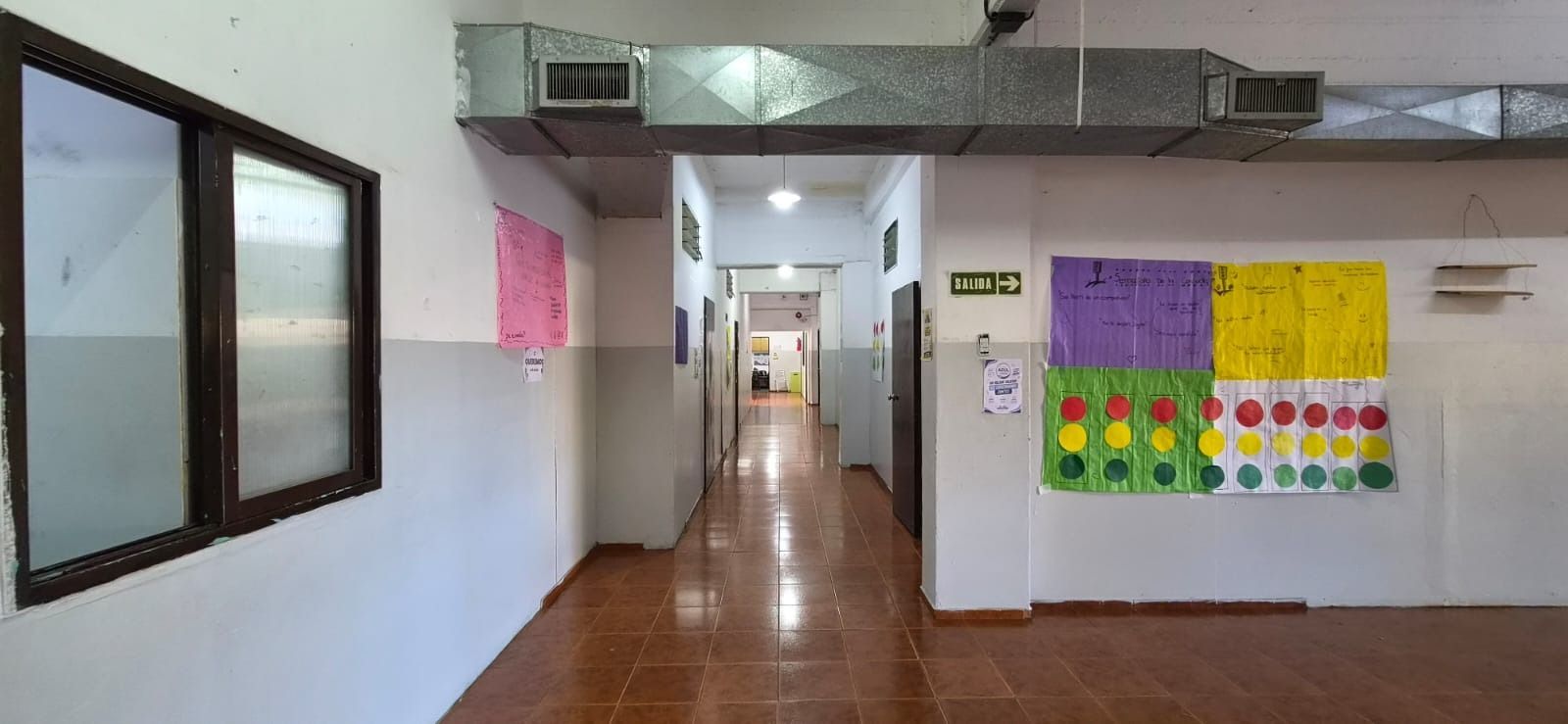 Pasillo escolar vacío con suelo brillante, ventanas a la izquierda y coloridos carteles en la pared derecha.