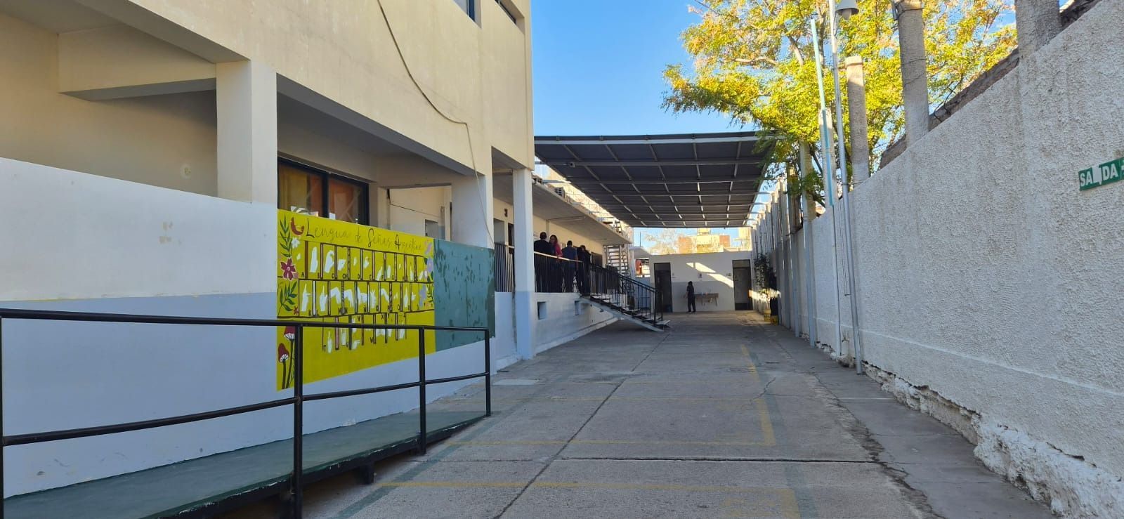 Un estrecho callejón de hormigón entre edificios, con una verja amarilla y árboles en la parte superior, que conduce a un luminoso patio.