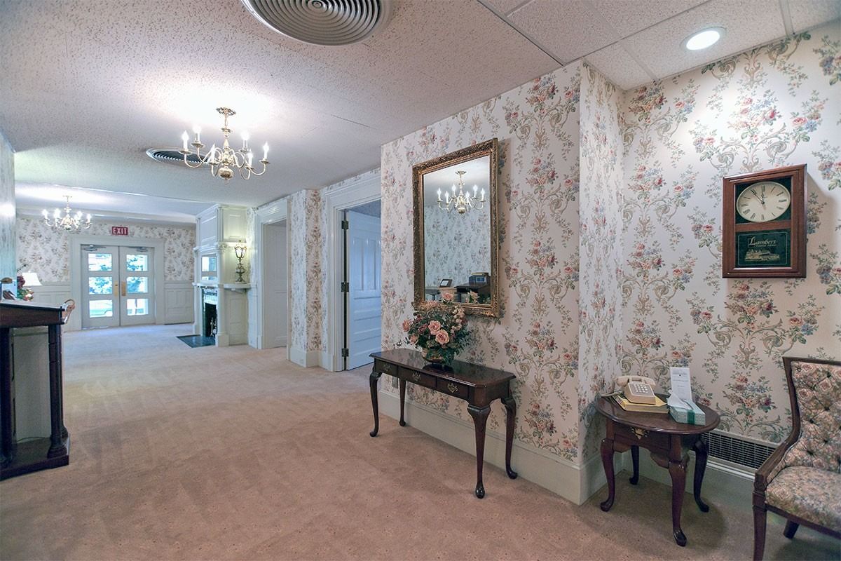 Elegant carpeted hallway with floral wallpaper, wall mirror, small tables, and chandelier lighting