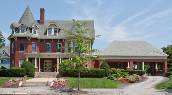 Large red-brick Victorian-style house with a covered driveway, manicured shrubs, and spring landscaping