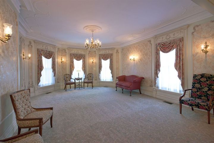 Elegant living room with floral wallpaper, chandeliers, red sofa, and chairs by large curtained windows