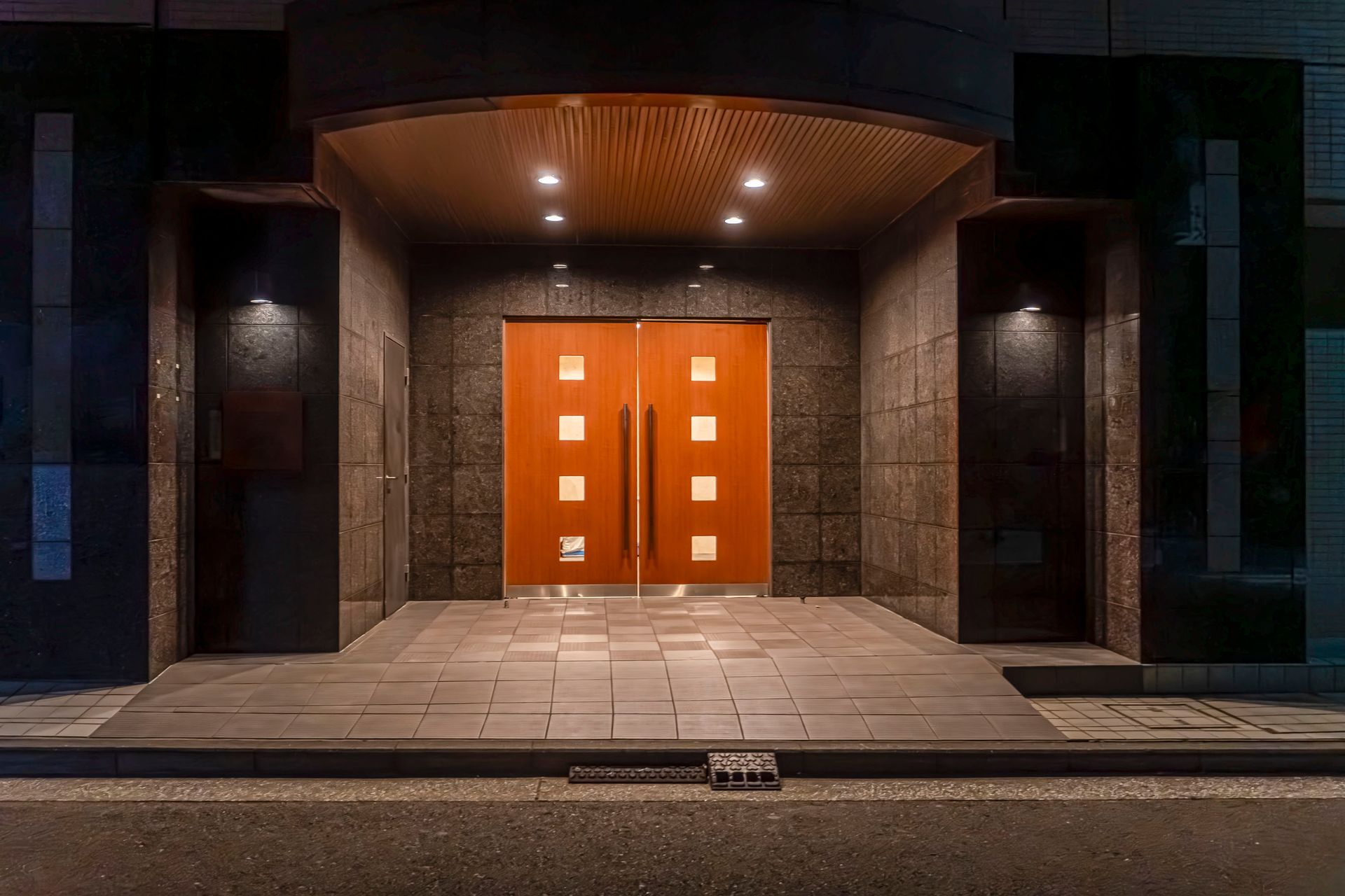 Exterior entrance with wooden doors, lit by overhead and side lights. Dark gray stone walls and a ramp.