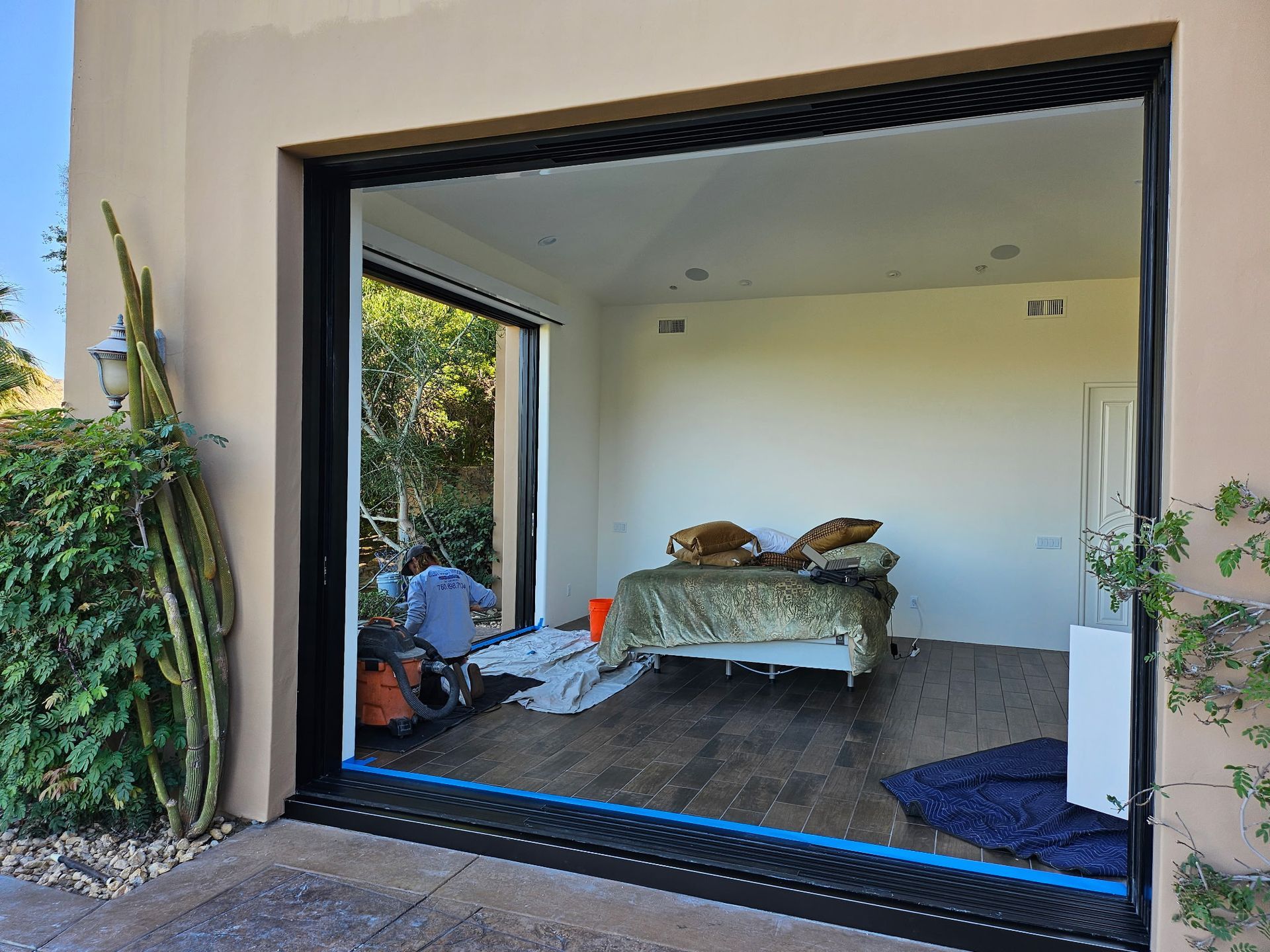 Open doorway frames interior room with bed. Outdoor view includes plants, patio, and blue painter's tape.