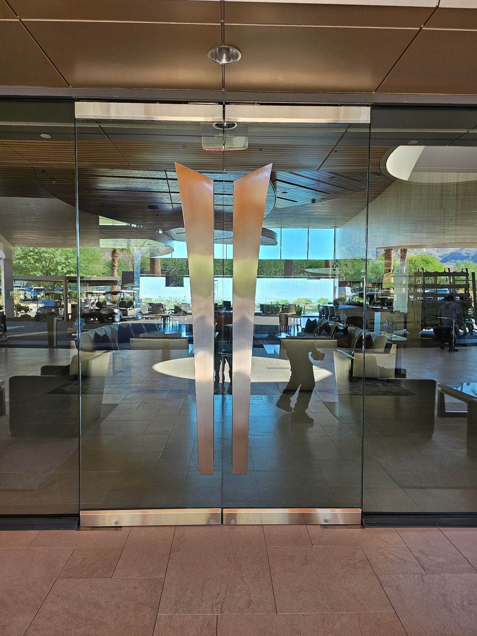 Glass doors with vertical metal handles. A person's reflection is visible inside. Natural light, indoor setting.