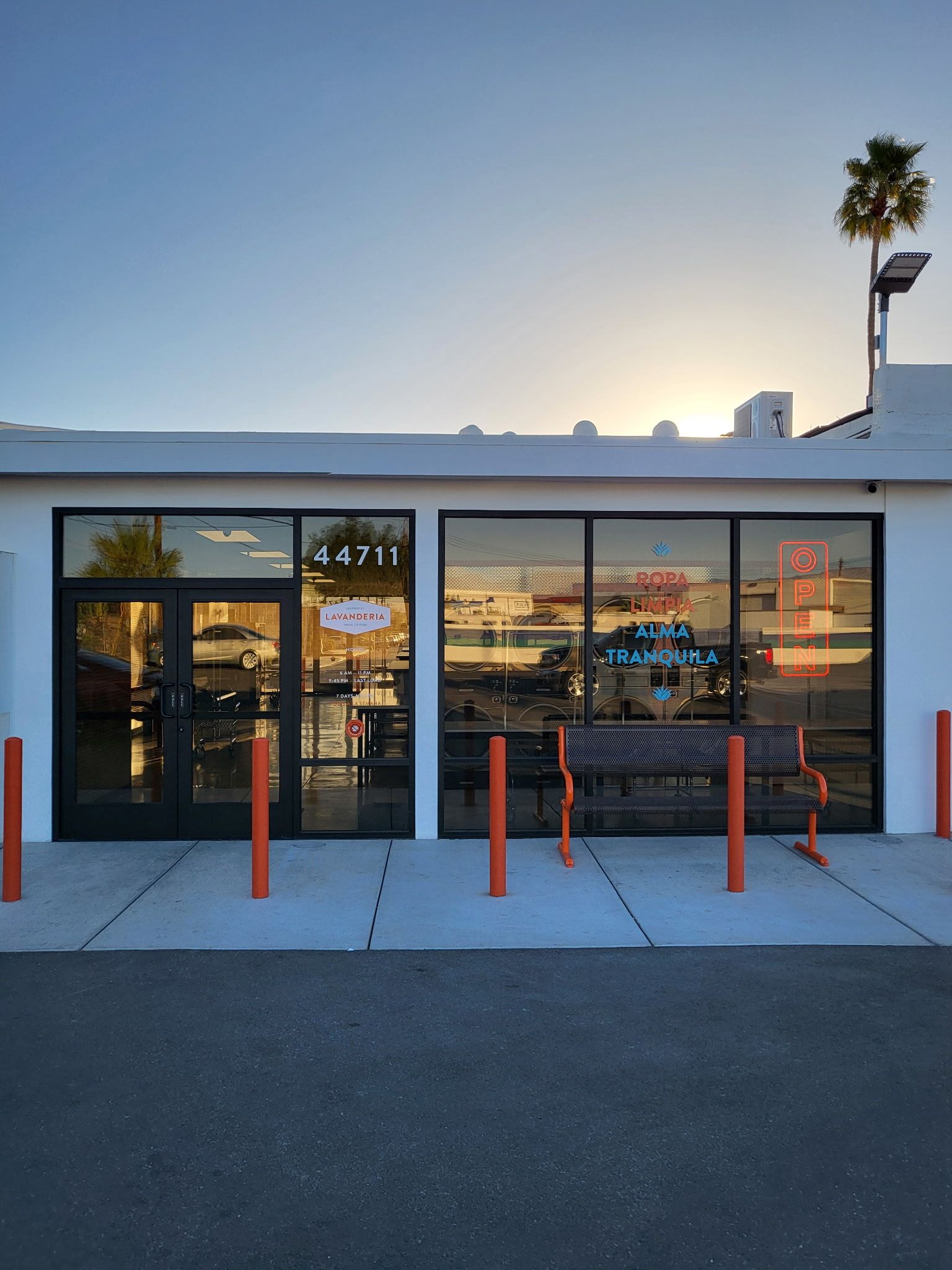 Exterior of a business with glass windows and door, address 44711. Orange bollards line the front. Sun setting in background.