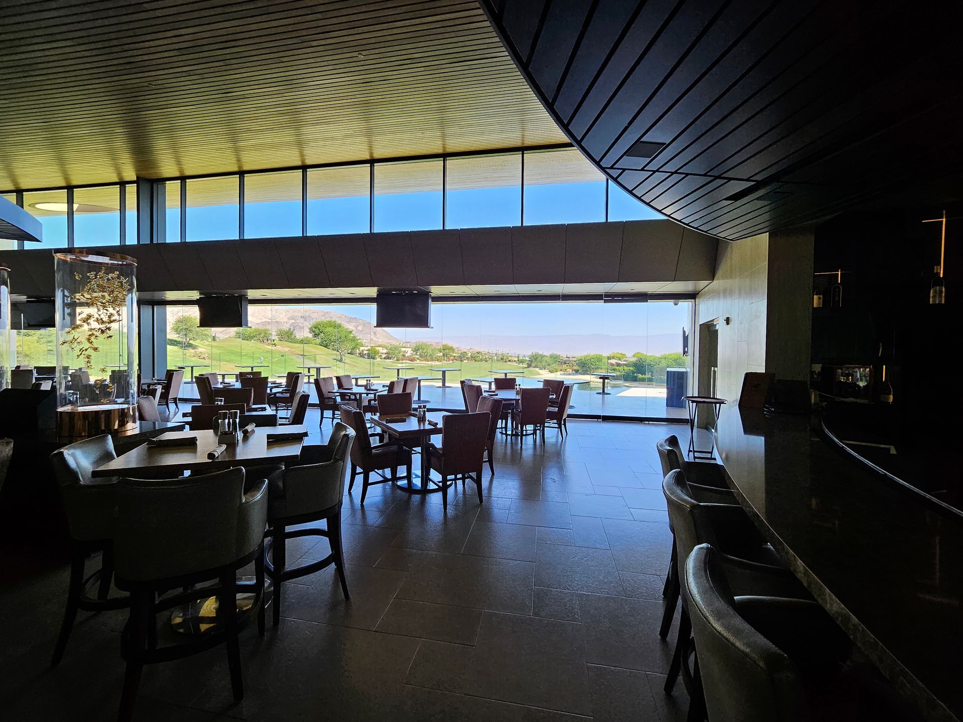 Interior of a restaurant with tables, chairs, and a bar overlooking a golf course.