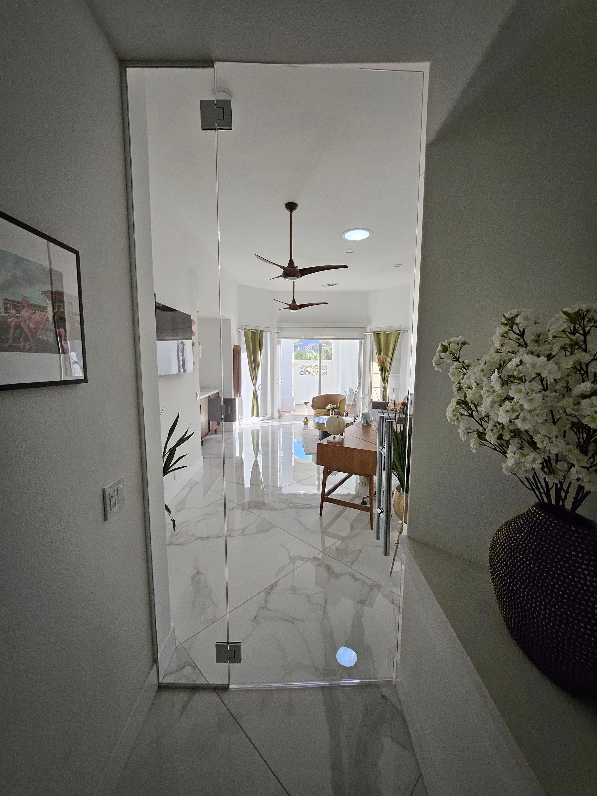 Hallway with glass door revealing a living room; white walls, marble floor, furniture, ceiling fan.