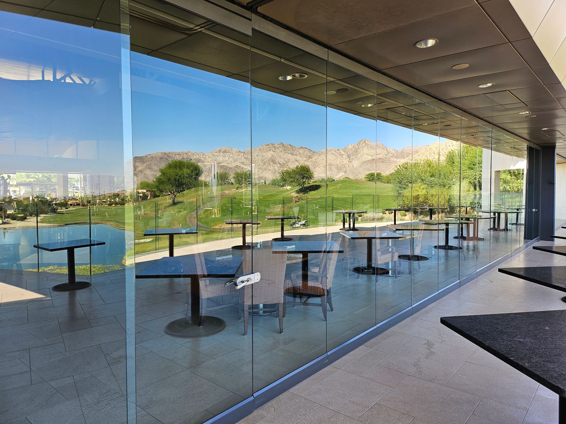 Glass-walled restaurant overlooking a golf course with tables and chairs, under a shaded overhang.