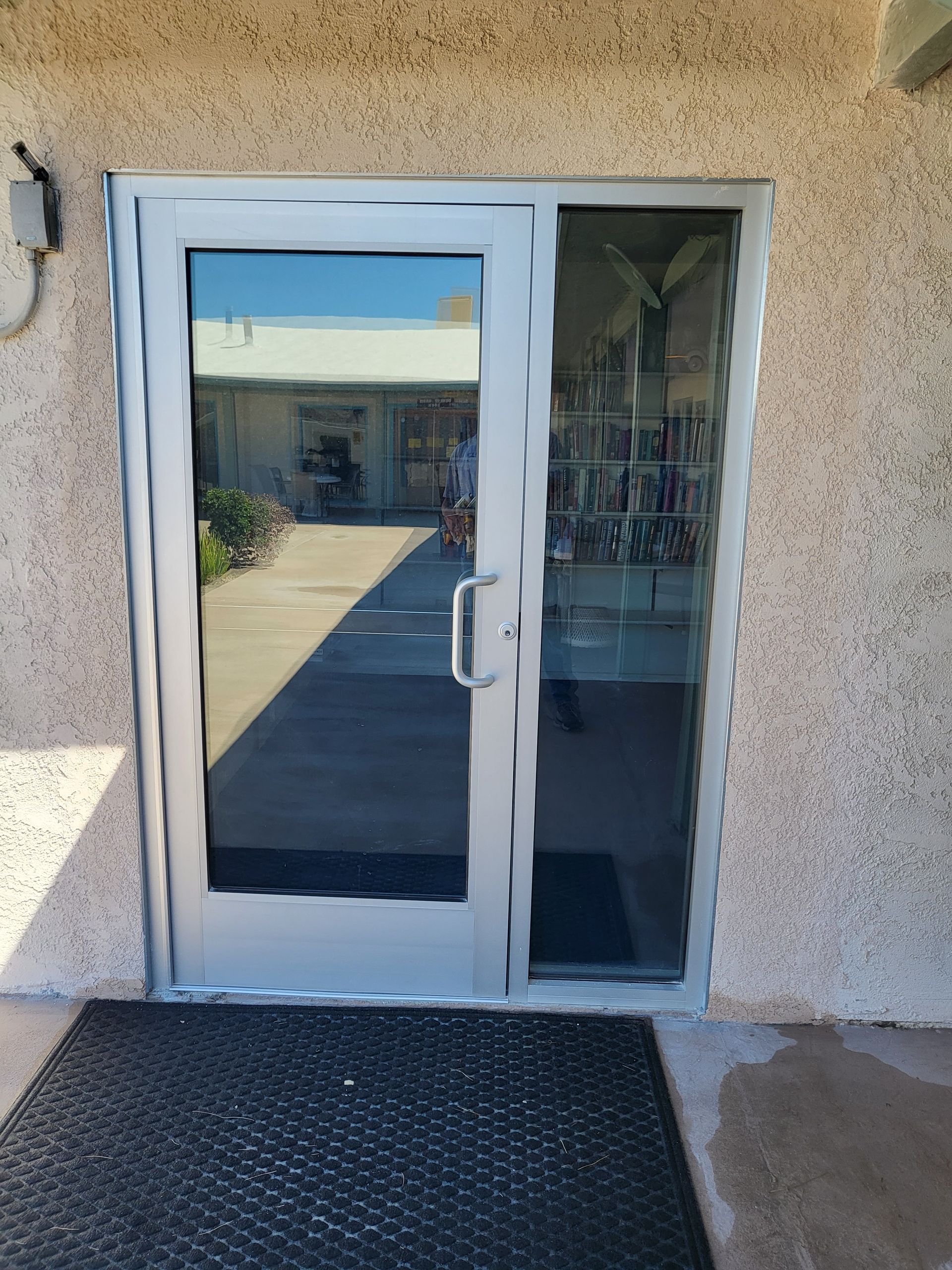 A silver-framed glass door with a sidelight, set in a stucco wall. A black mat is at the entrance.
