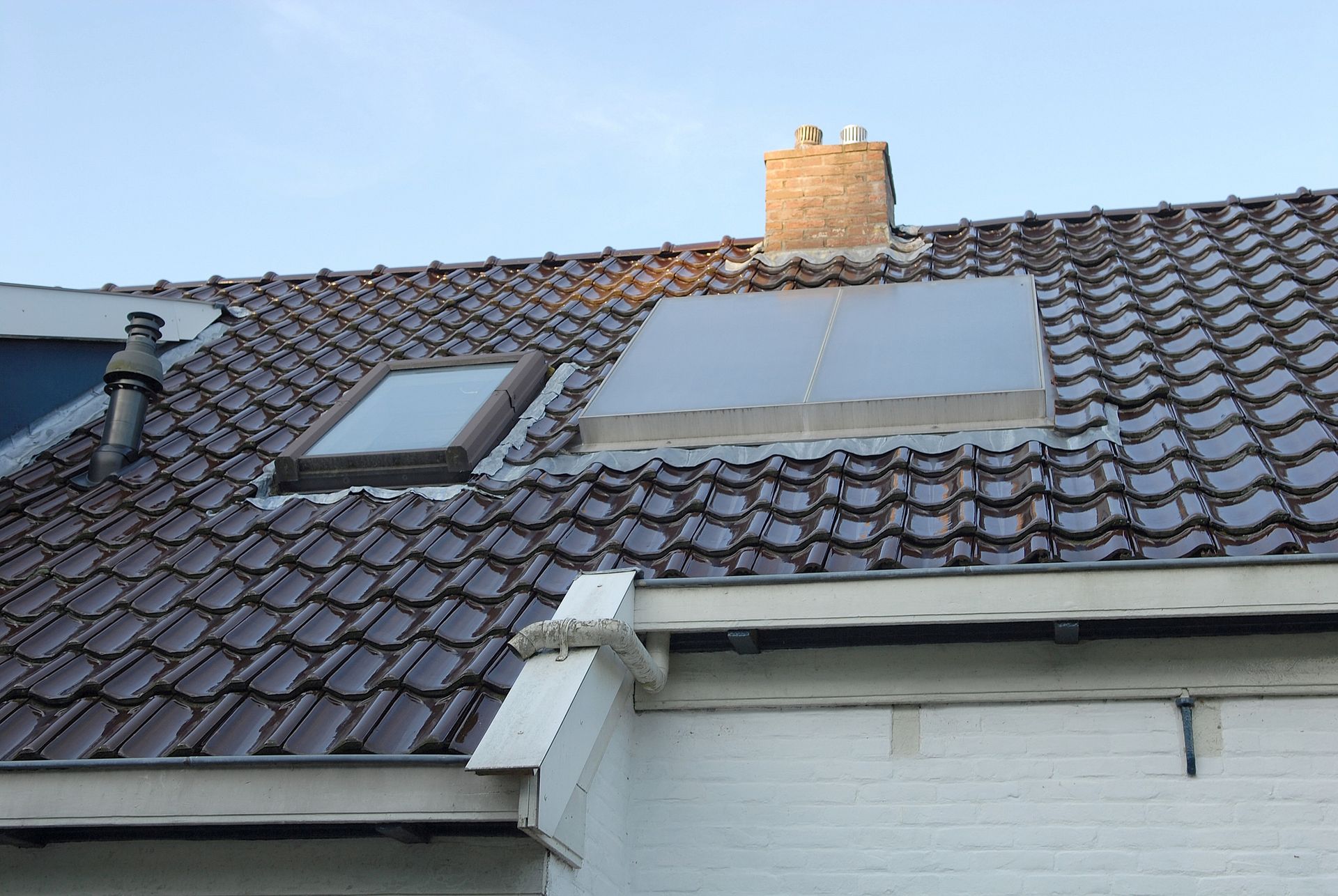 Brown tiled roof with two skylights and a brick chimney under a blue sky.