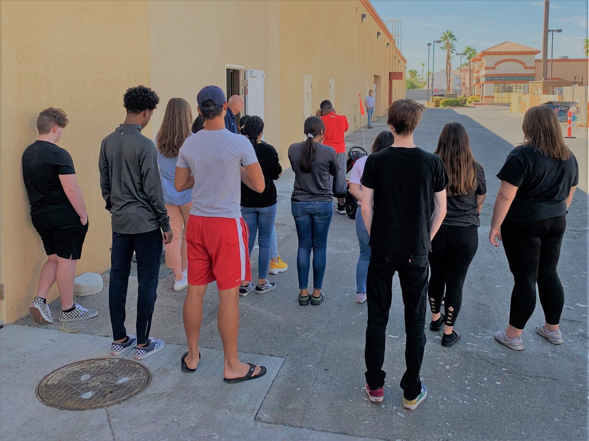 Small crowd standing outdoors in an alley beside a beige building, facing away and looking ahead.