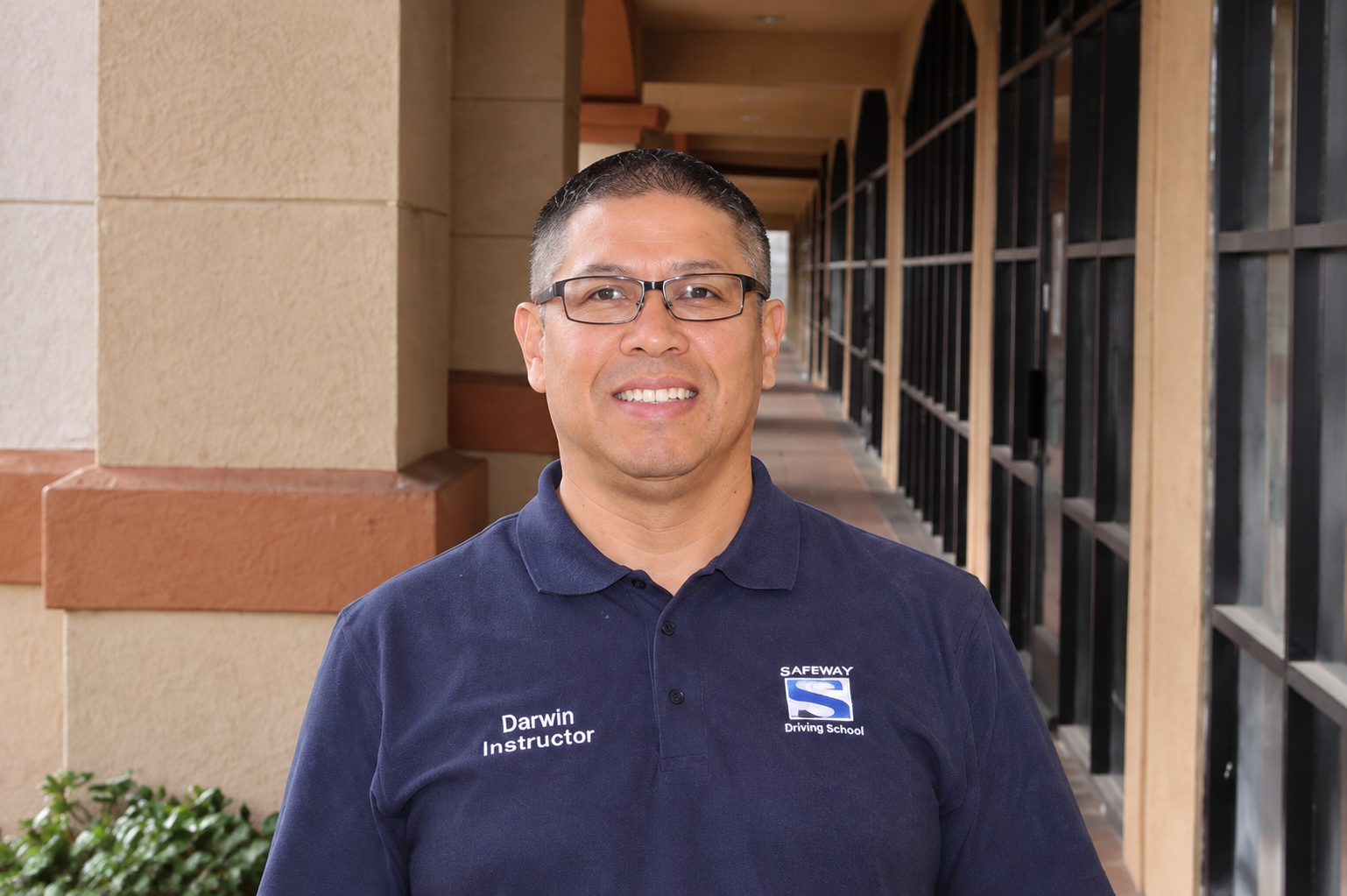 Man standing outdoors in front of a building, wearing glasses and a navy polo shirt, smiling.