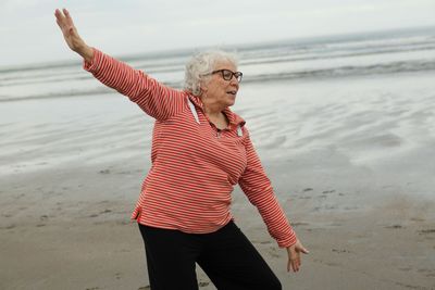 Woman in red striped shirt and black pants doing Tai Chi on a beach near the ocean.