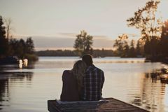 Couple sits on a dock, backs to the viewer, overlooking a lake at sunset.