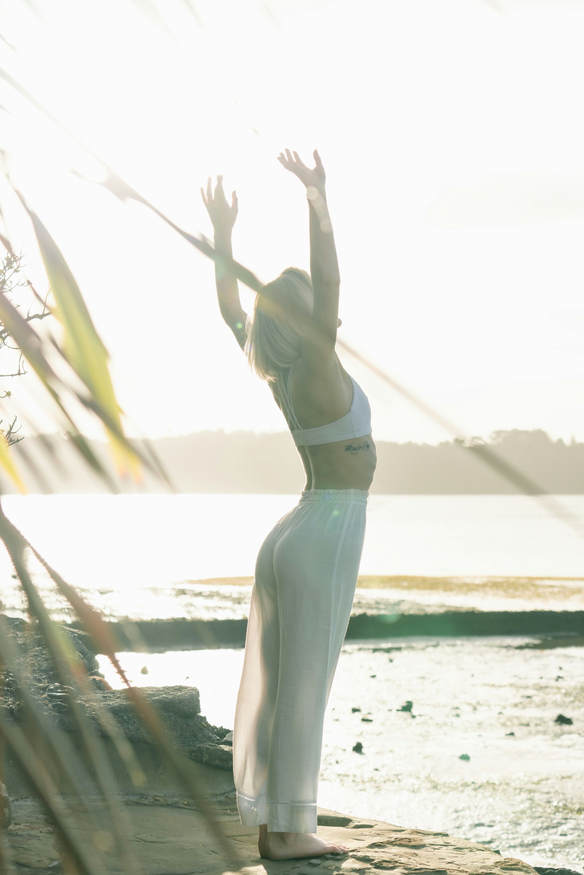 Person standing on a beach with arms raised at sunset, silhouetted against bright water.