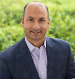 Headshot of a smiling man in a blue blazer against a blurred green outdoor background