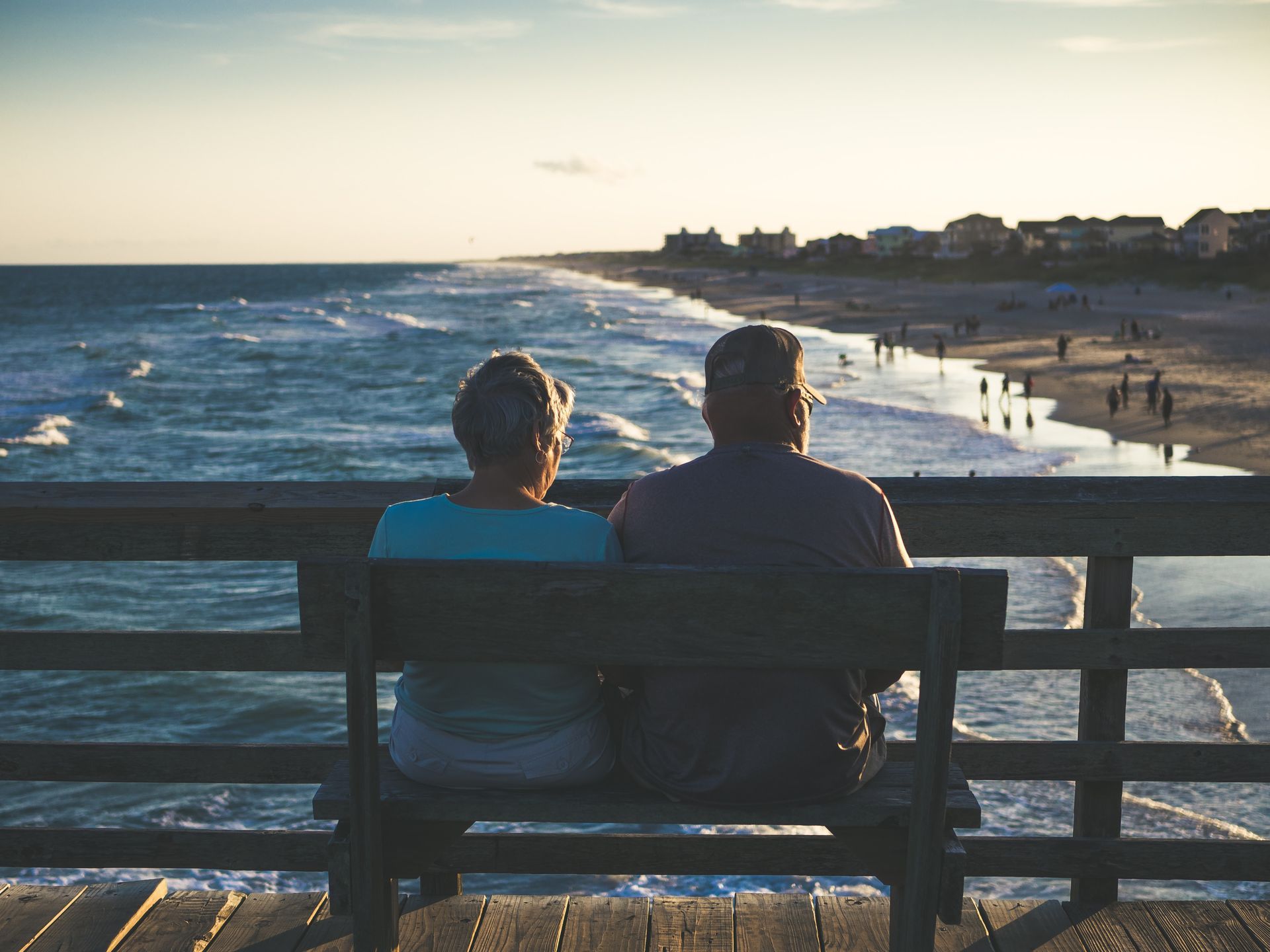 Two people sit on a bench overlooking the ocean at sunset, with waves and a sandy beach in view.