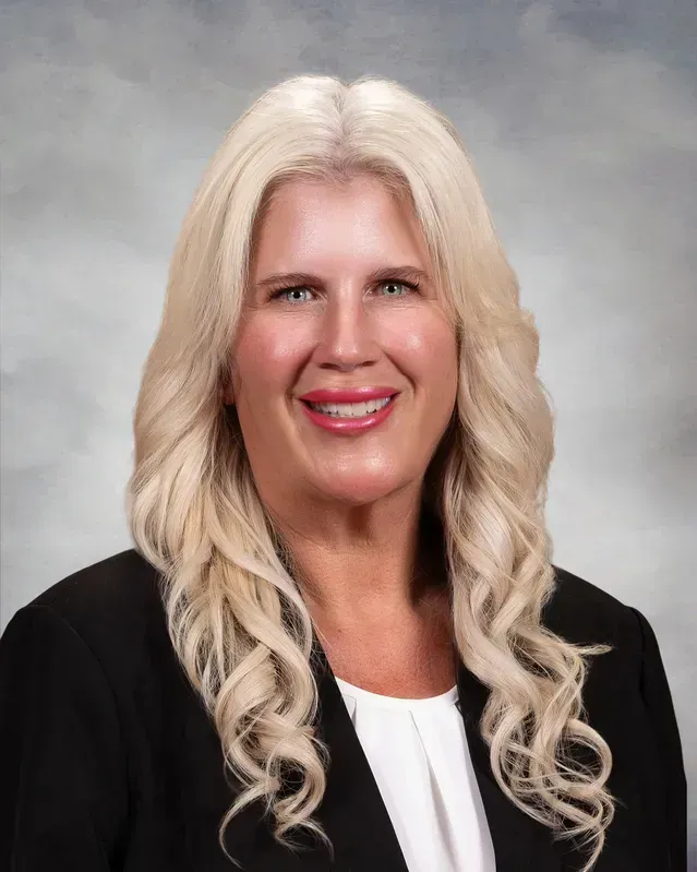 Woman with long blonde wavy hair, wearing a black blazer and white top, smiling, headshot.