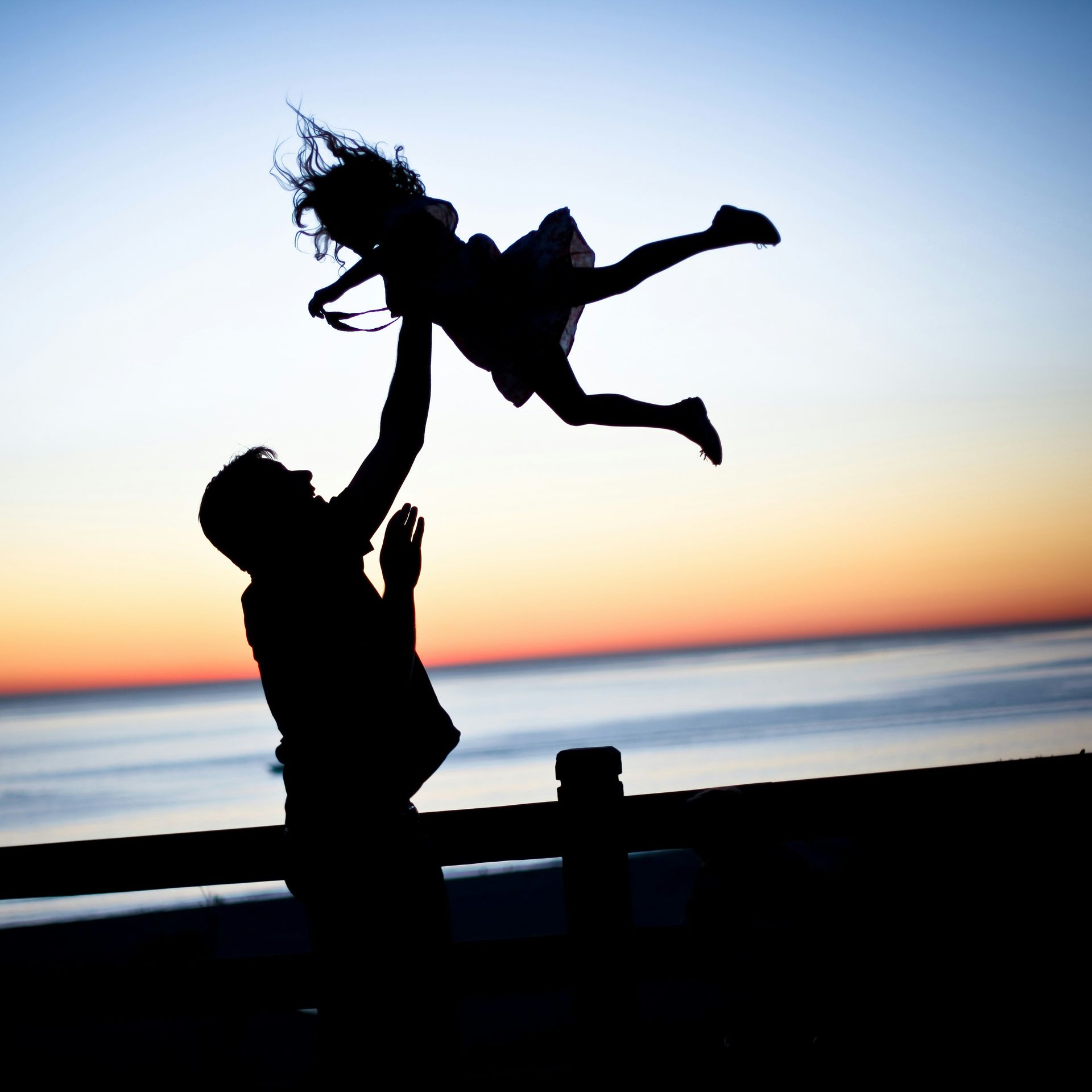 Silhouetted adult lifting child overhead at sunset by the sea