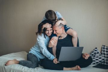 Family of three on a bed, smiling at a laptop. Child on father's shoulders, mother hugging.