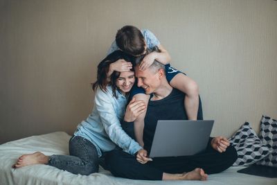 Family on bed; child on dad's shoulders, mom embraces dad, all smiling while dad uses laptop.