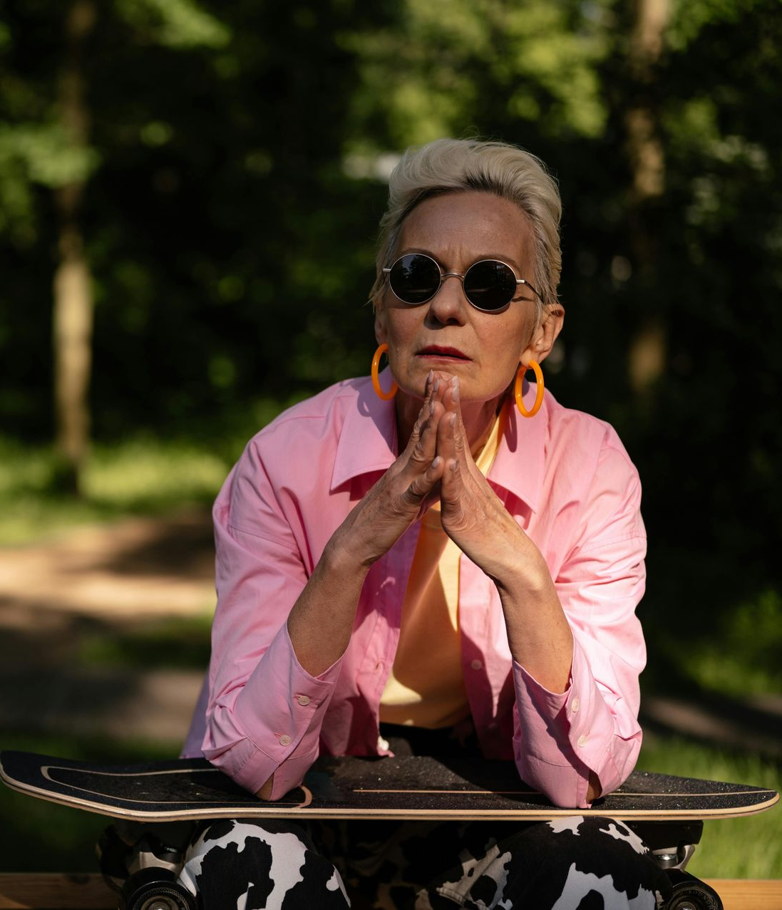 Person in pink shirt and sunglasses leaning on a skateboard outdoors, hands clasped under chin.