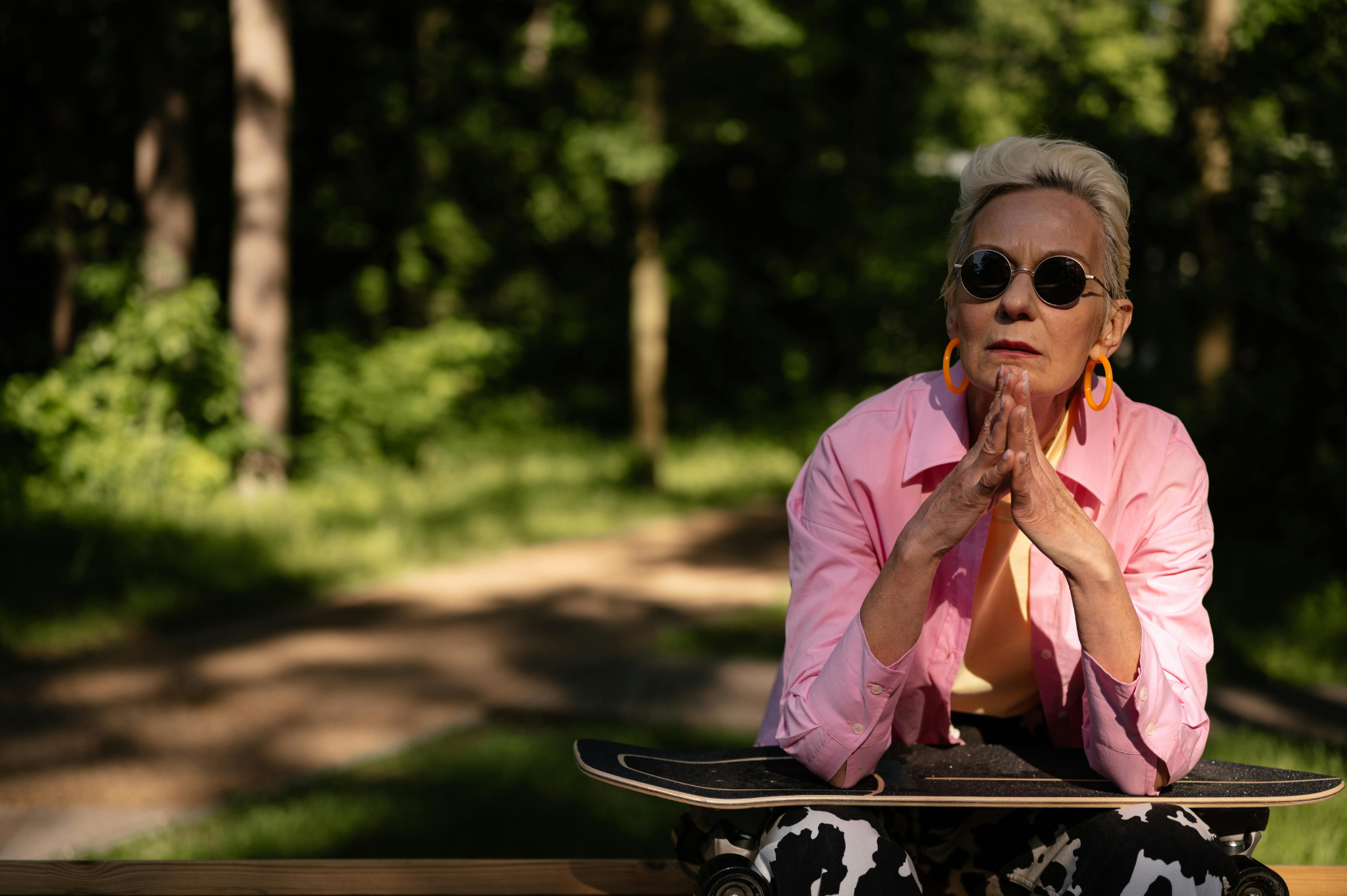 Person in pink jacket and sunglasses sits on skateboard, hands clasped, outdoors.