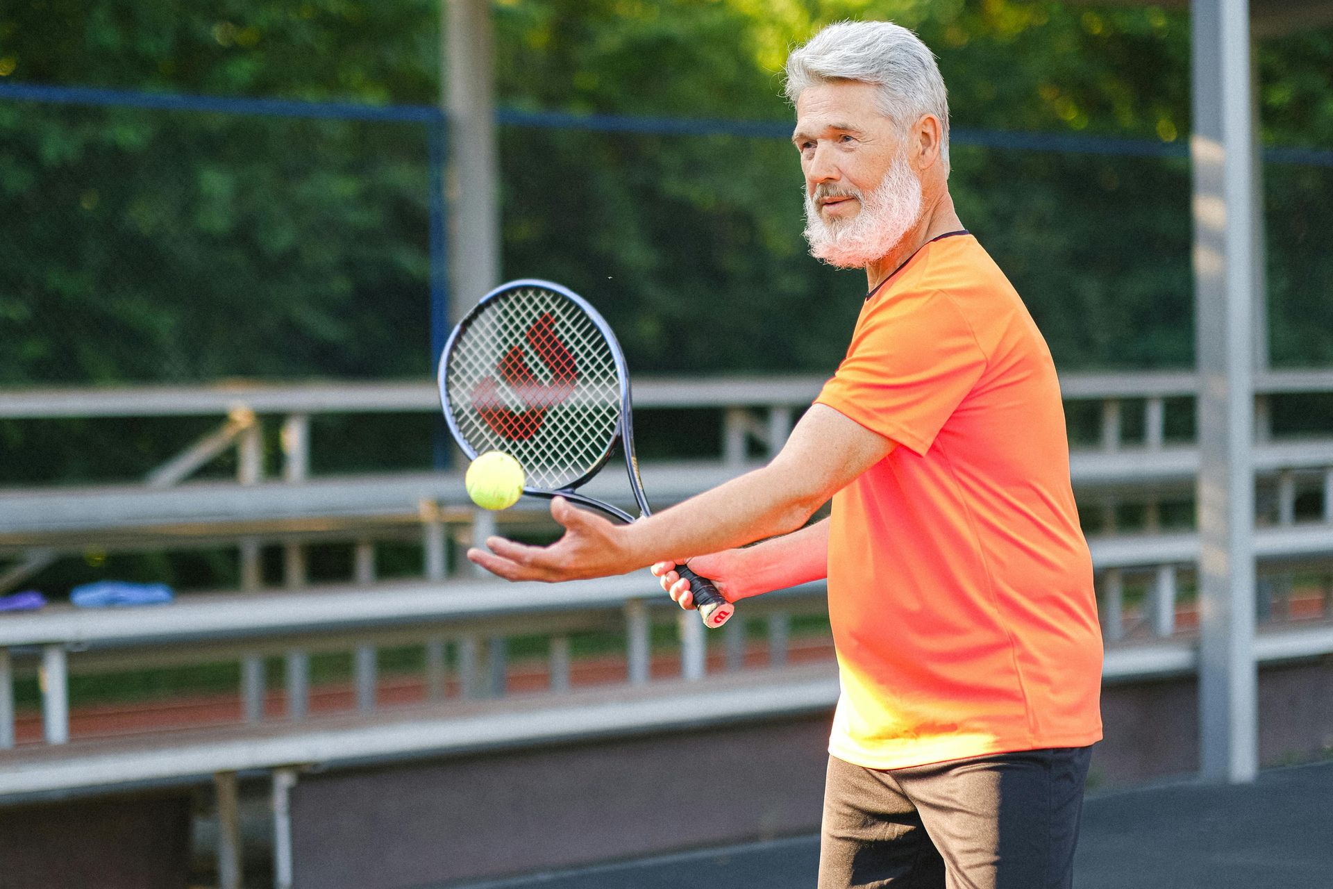 Man in orange shirt tossing tennis ball on court, holding racket.