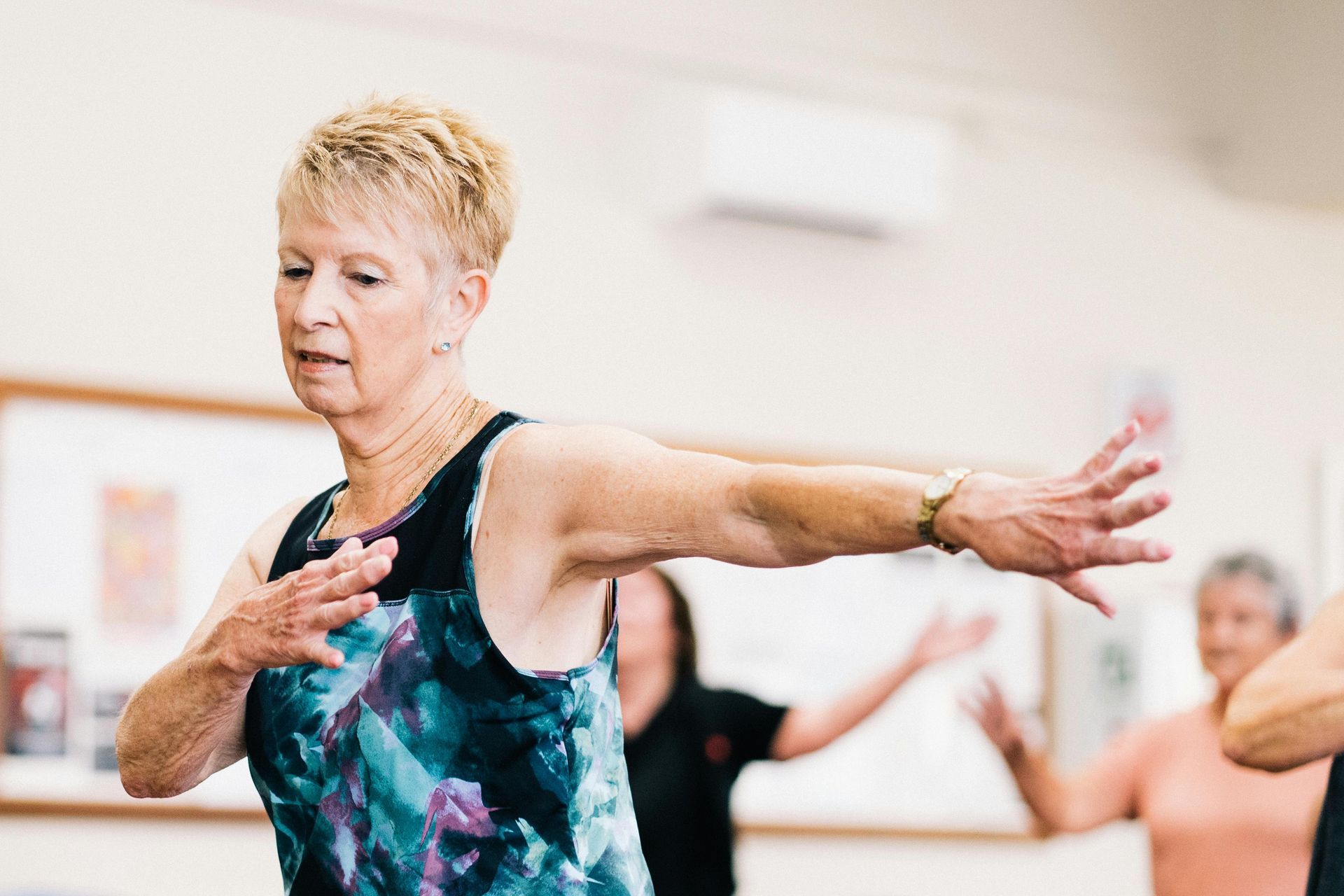Woman in exercise class, extending arm. Others in background. Indoors.