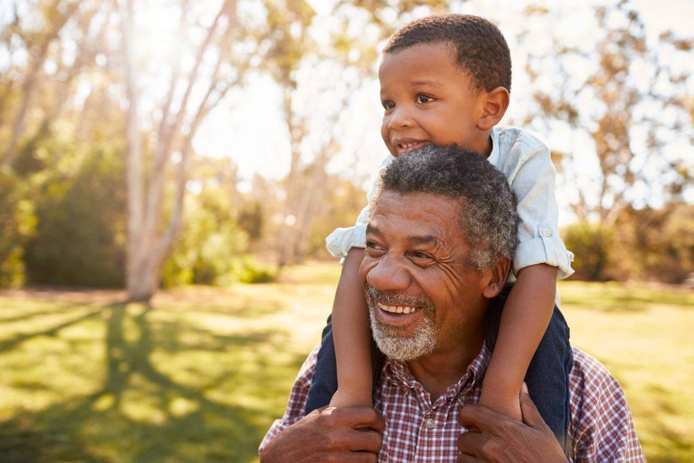 Grandfather carrying child on shoulders in sunny park; both smiling.