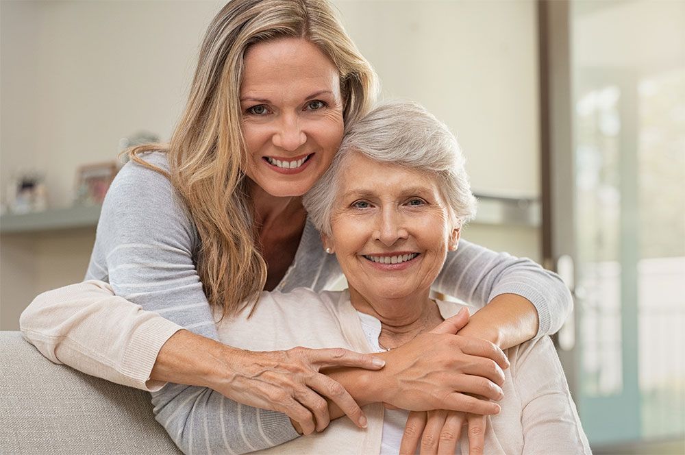 Woman embraces elderly woman, both smiling, indoors.