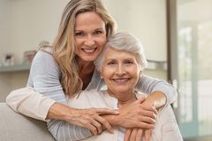 Woman embracing an older woman; both smiling indoors.