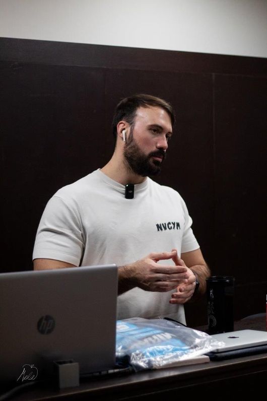 Nicola si allena in palestra, mostrando il suo fisico muscoloso. Sorride con le braccia piegate, mettendo in mostra i bicipiti.