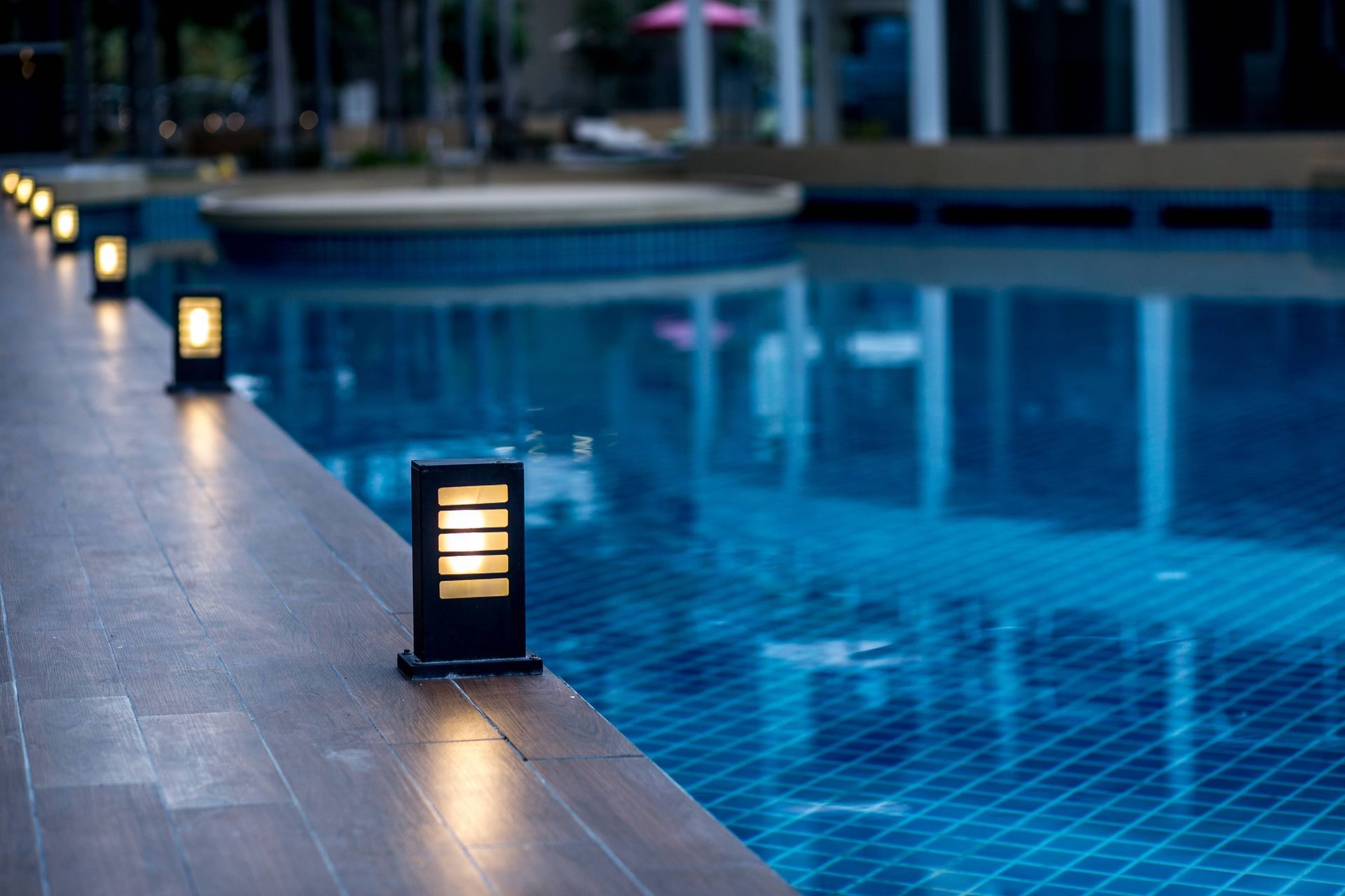 Poolside lights illuminate a wooden deck next to a blue swimming pool.