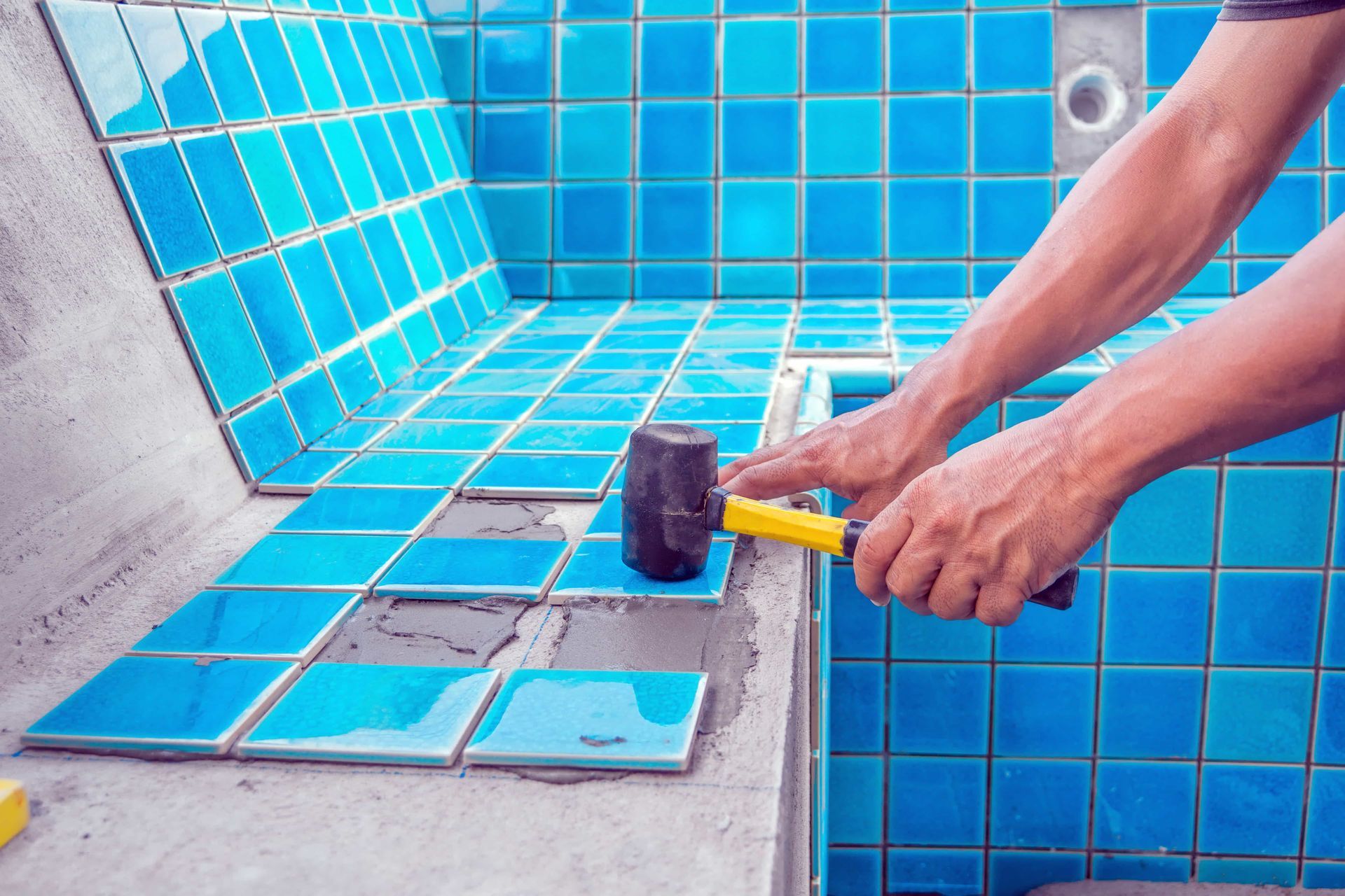 Person tiling a pool with blue tiles, using a rubber mallet.