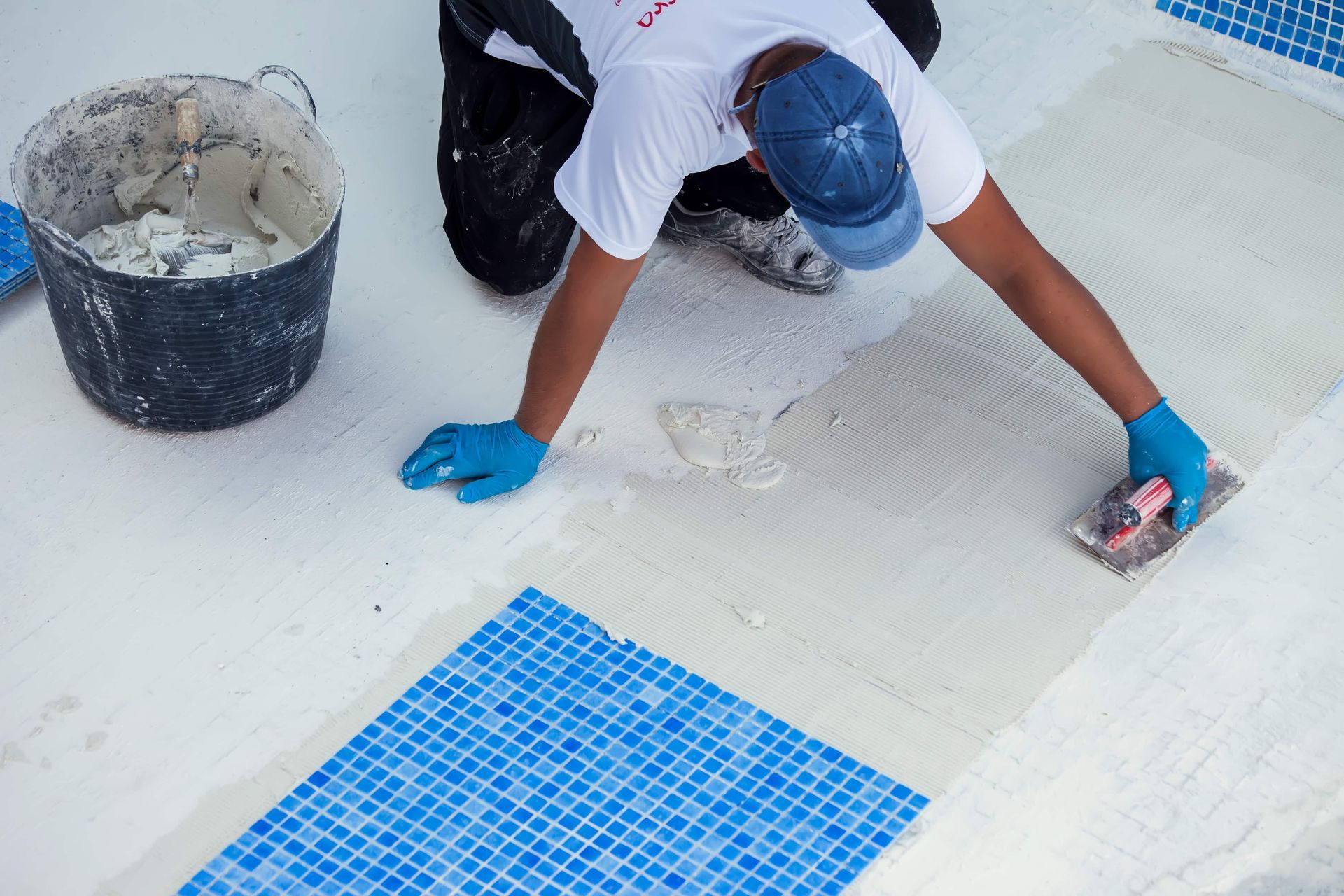 Person tiling a pool, applying mortar with a trowel, wearing blue gloves and cap.