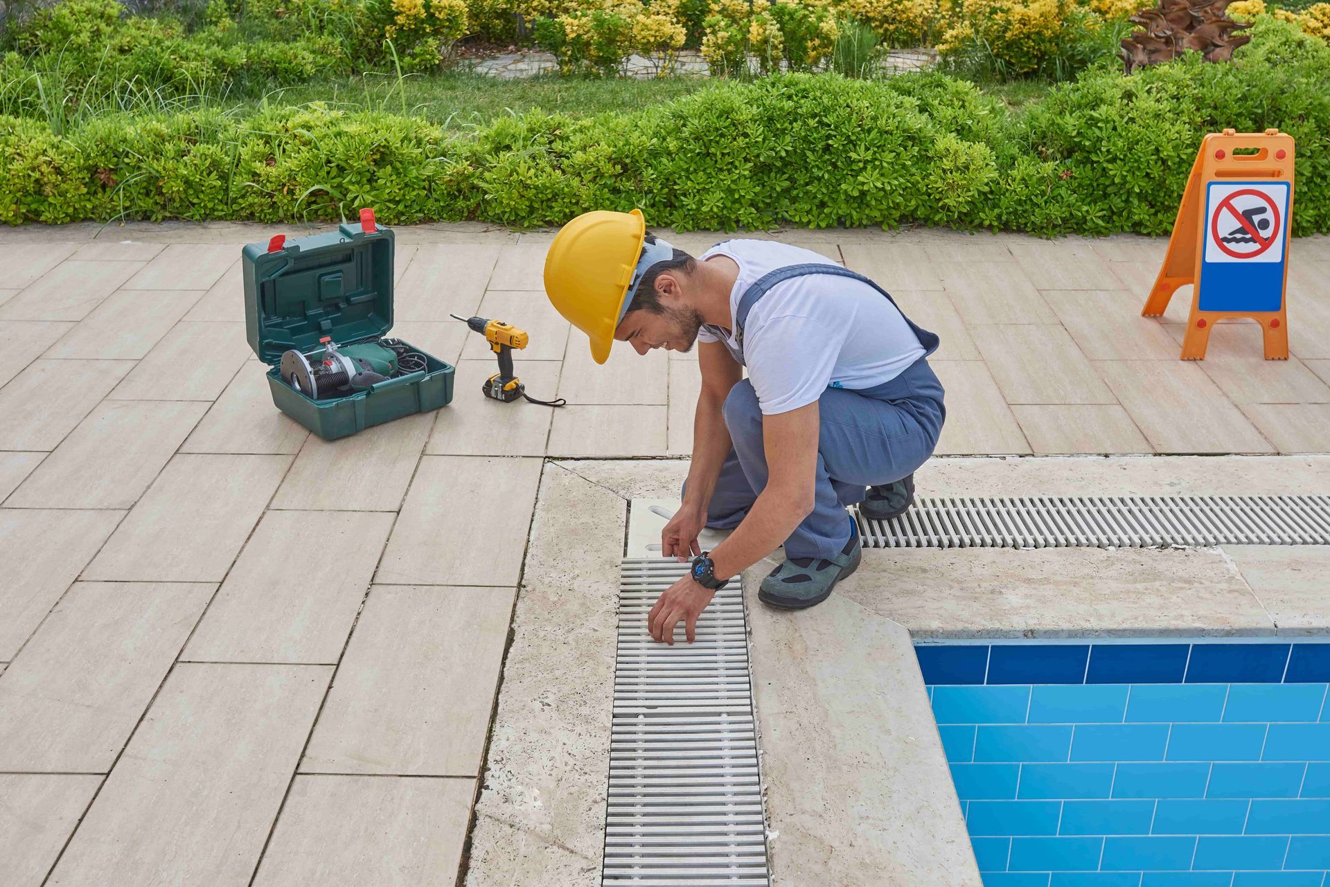 Pool technician in overalls and hard hat fixing a pool drain, tools nearby.
