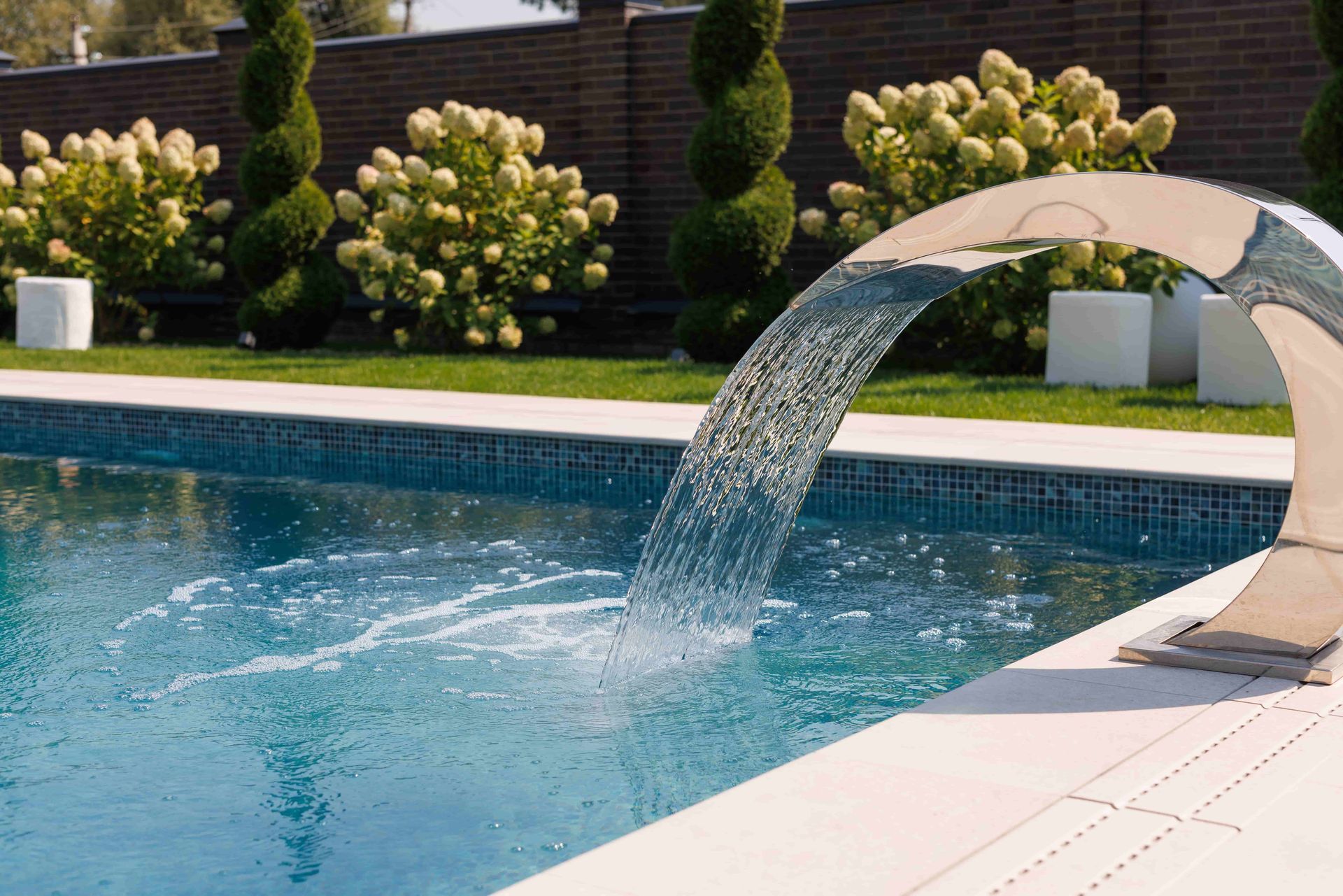 Pool with a chrome waterfall feature; green grass and shrubs in the background.