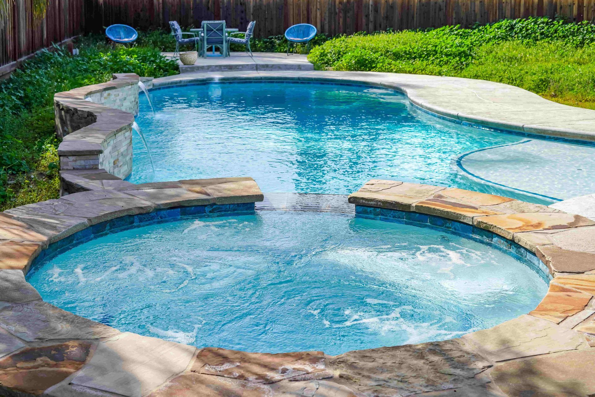 A blue pool and hot tub with a waterfall feature in a backyard with outdoor seating.
