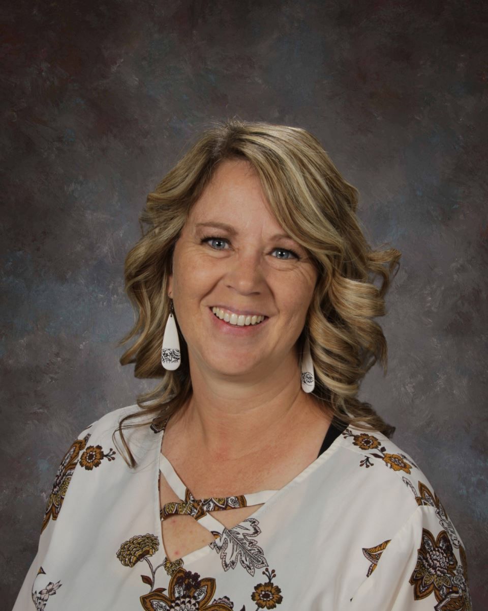 A woman wearing a white floral shirt and earrings is smiling for the camera.