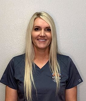 A woman with long blonde hair is wearing a gray scrub top and smiling.