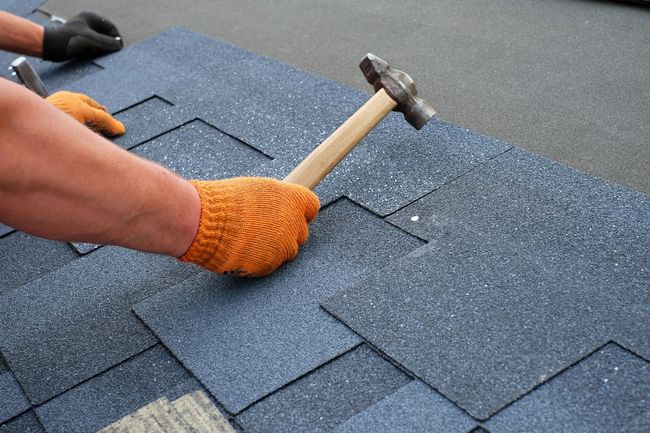 Worker hammering shingles on a roof with orange gloves and a black glove nearby Worker hammering shingles on a roof with orange gloves and a black glove nearby