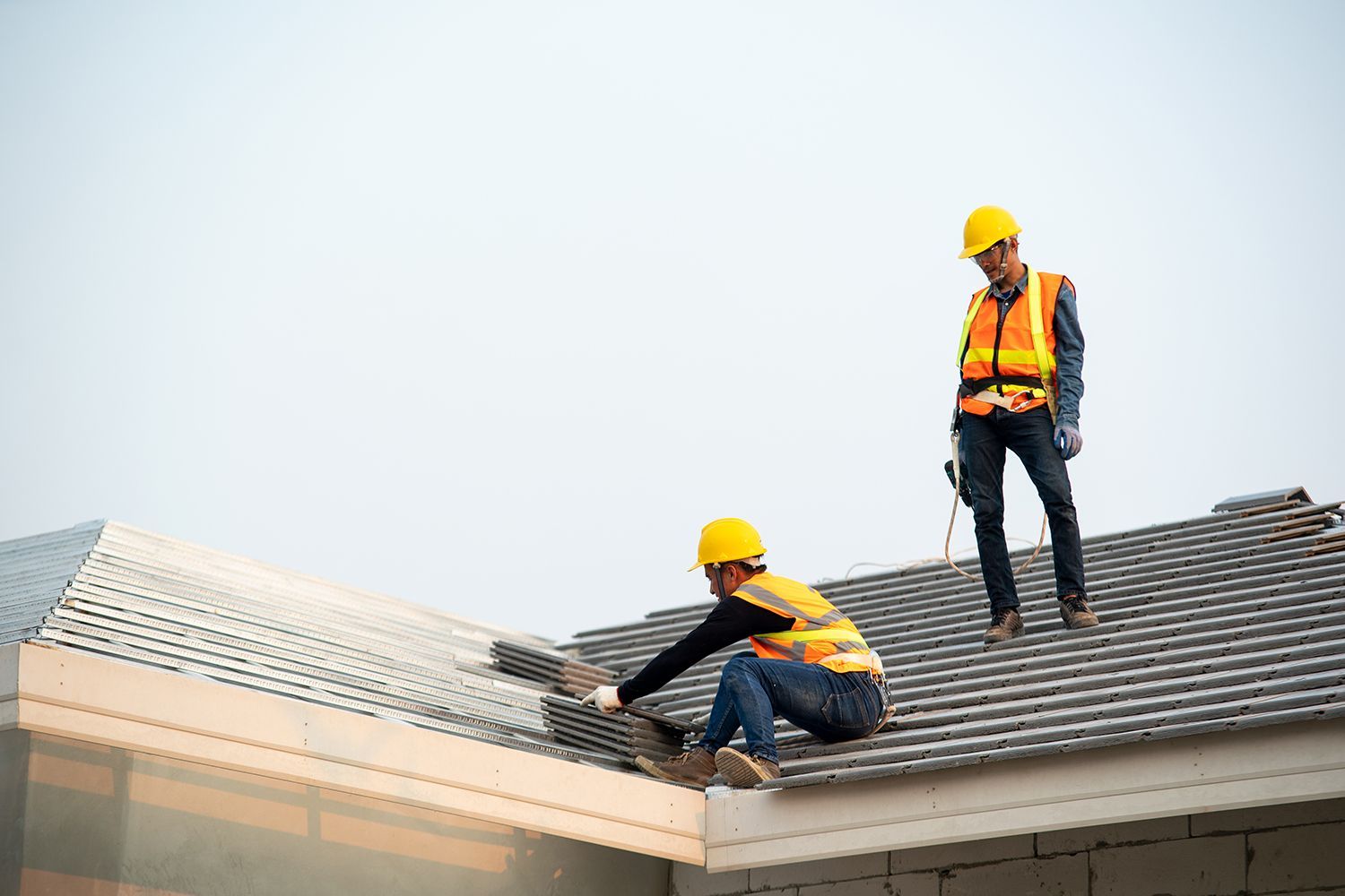 Two roofers in hard hats working on a house roof, one kneeling and one standing with safety harnesses Two roofers in hard hats working on a house roof, one kneeling and one standing with safety harnesses