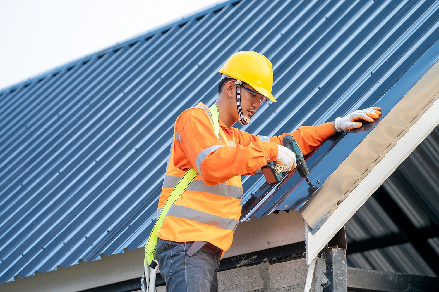 Worker in orange safety gear repairing a blue metal roof with a power drill Worker in orange safety gear repairing a blue metal roof with a power drill