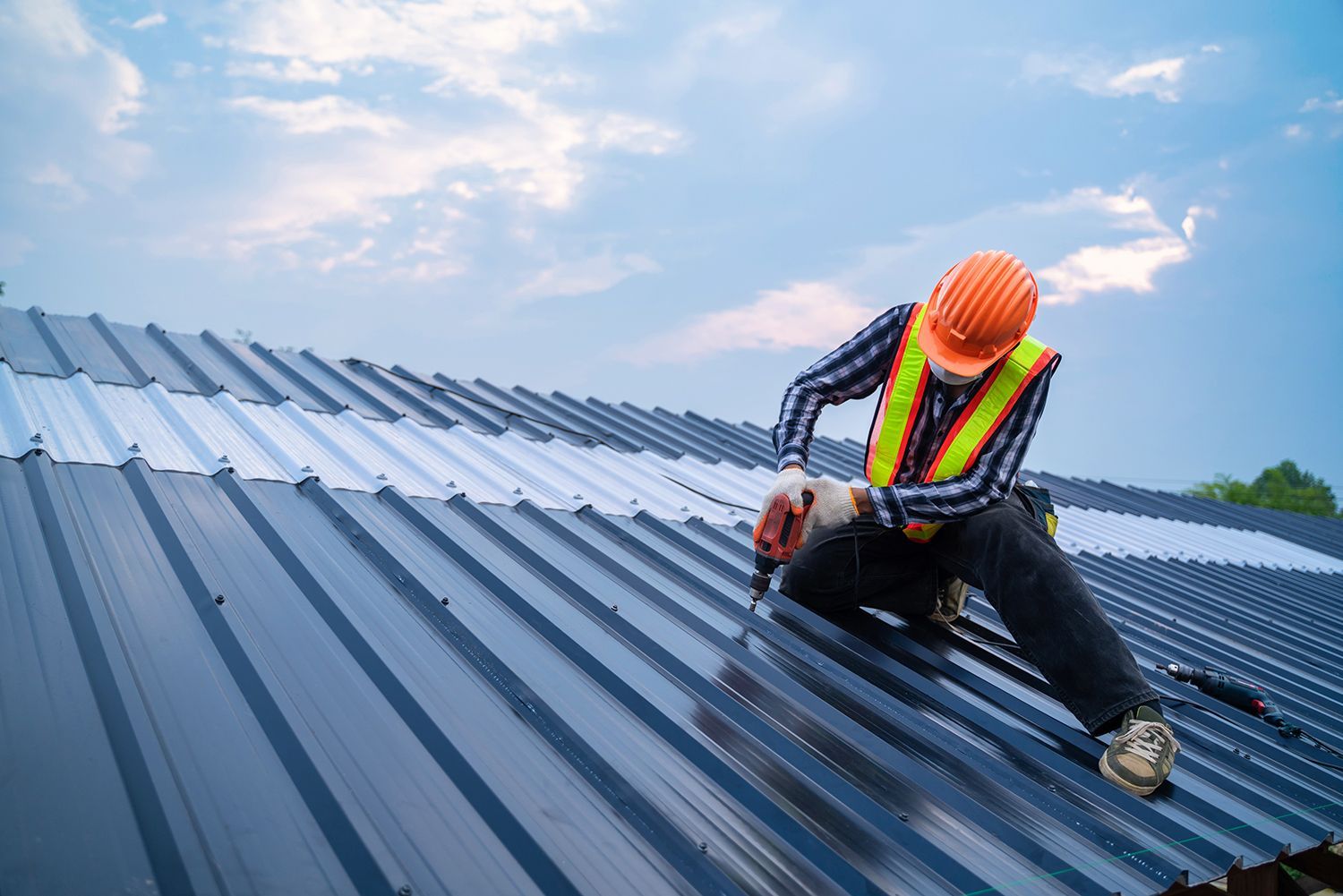 Worker in orange hard hat and safety vest securing blue metal roofing on a rooftop under a partly cloudy sky Worker in orange hard hat and safety vest securing blue metal roofing on a rooftop under a partly cloudy sky