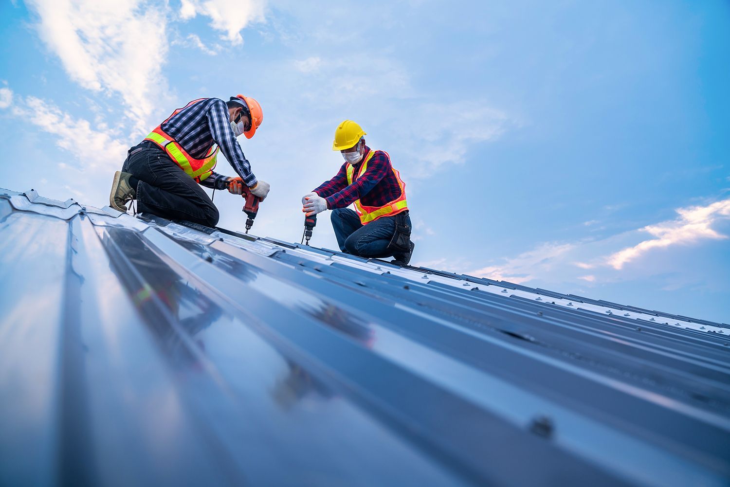 Two roofers in hard hats installing metal roofing on a steep roof under a blue sky Two roofers in hard hats installing metal roofing on a steep roof under a blue sky