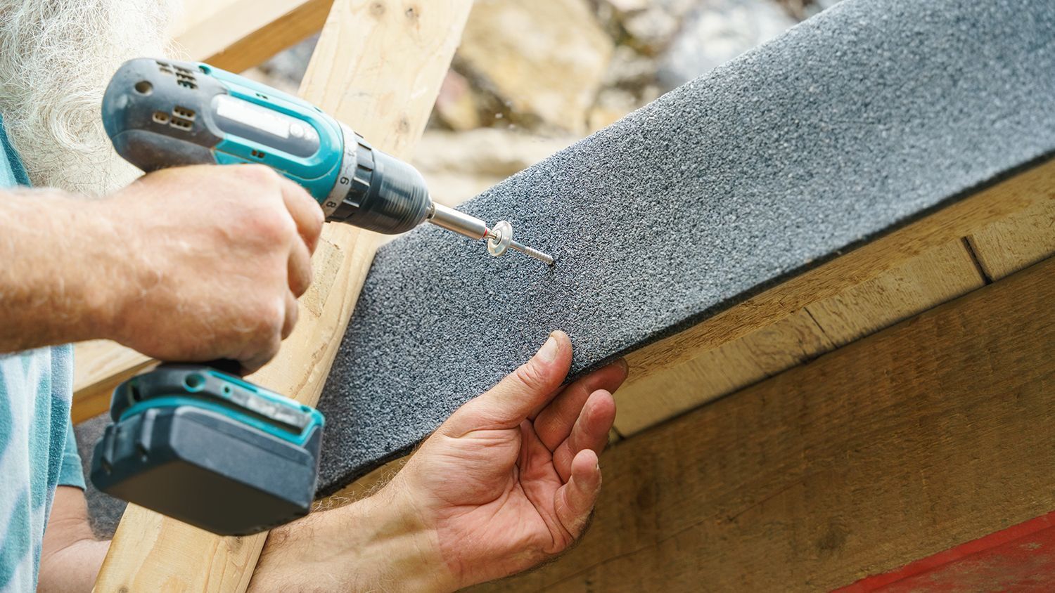 Hands using a cordless drill to fasten gray roofing shingles on a roof edge.
