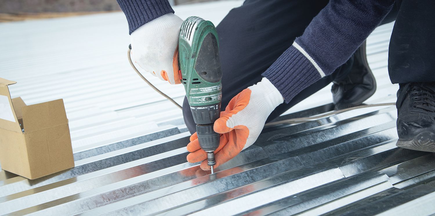 Hands using a green drill to fasten a corrugated metal roof panel outdoors Hands using a green drill to fasten a corrugated metal roof panel outdoors